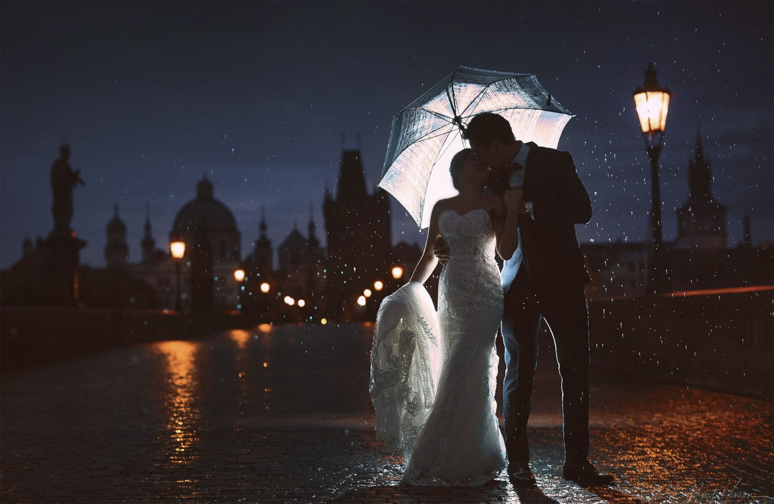 As the rain pours around them, a bride & groom are silhouetted against the night skyline of the Charles Bridge in Prague.