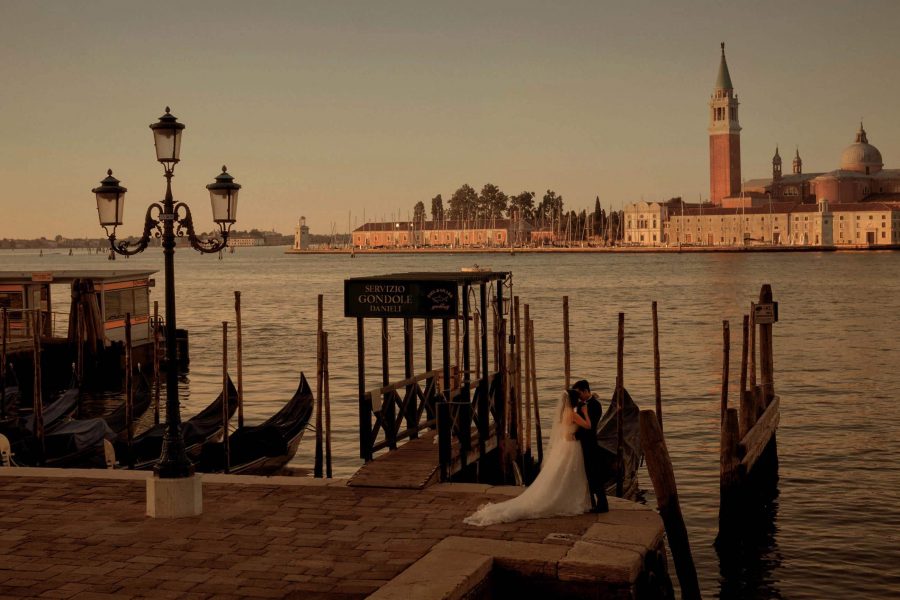 A bride & groom embrace in a deserted landscape near the Gondolas as the sunrise illuminates the Church of San Giorgio Maggiore across the lagoon in Venice Italy. Venice portrait photography by Kurt Vinion.