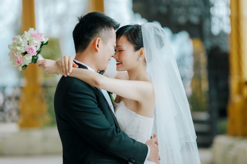 A smiling Asian bride lovingly embraces her groom as she clutches her bouquet behind his head.