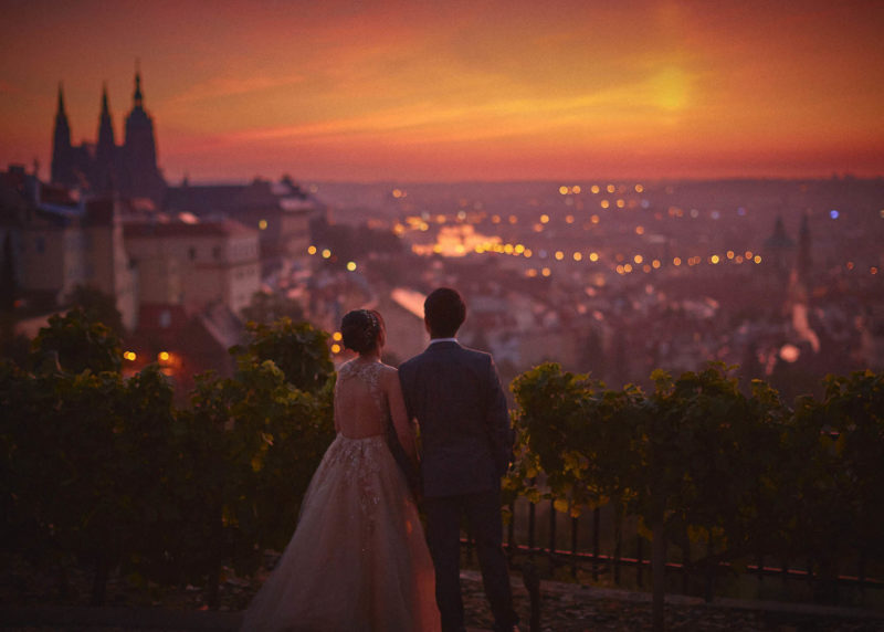 A stylishly dressed couple, whose arms are wrapped around each other, take in the landscape of Prague from above the castle as the sun rises.