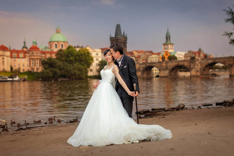 An elegantly dressed bride and groom, who is holding a walking stick, share a loving embrace near the river as the Charles Bridge dominates their background.