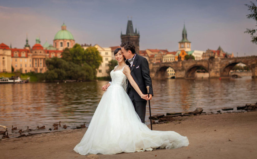 A stylish bride is lovingly embraced by her groom as he holds onto an antique walking stick near the river as the Charles Bridge dominates their background.