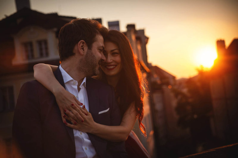 A smiling woman wraps her arms around her partner as the sun breaks above the skyline behind them.