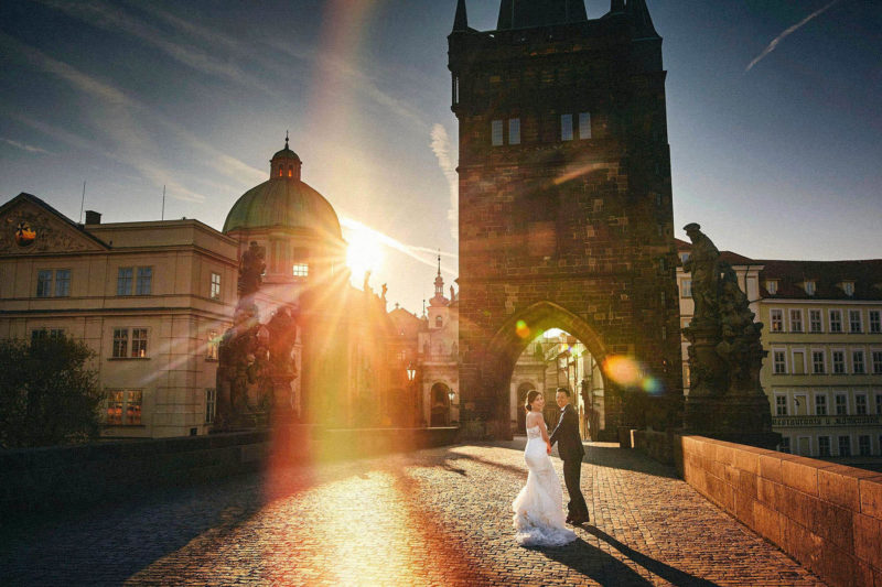 A bride is led by her groom as they run towards the sun-flared gothic tower on the Charles Bridge.