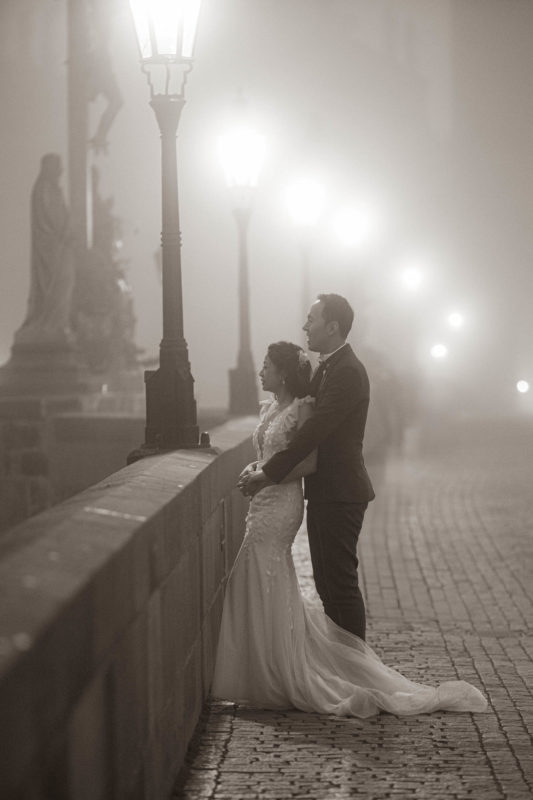 A monochrome image of a stylishly dressed couple embracing near the Calvary statue during their foggy Prague pre-wedding photo session.