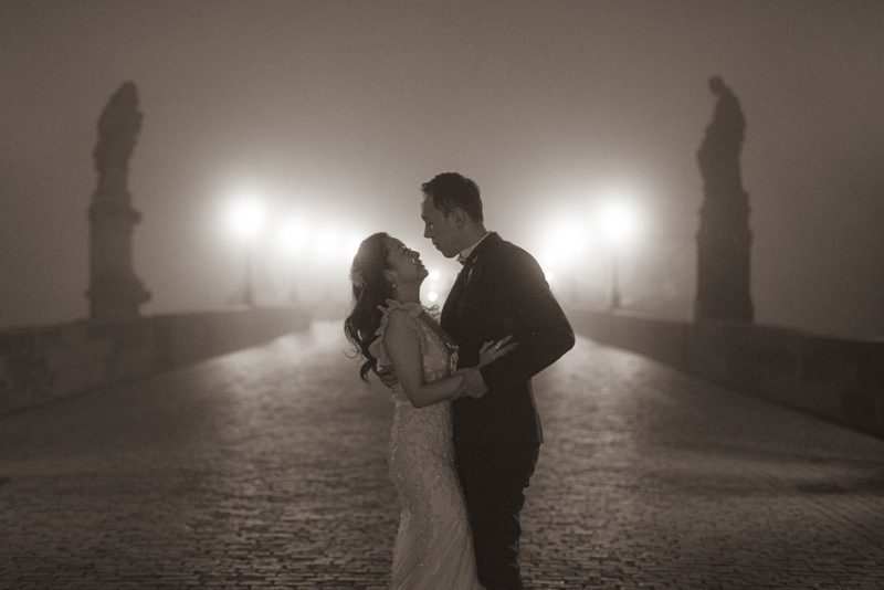 An elegantly dressed couple, embrace in the fog atop the Charles Bridge at night.