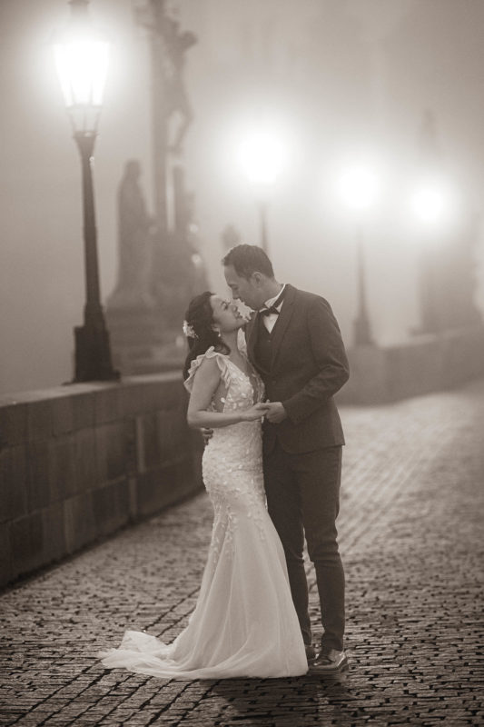 A monochrome image of a stylishly dressed couple embracing near the fog shrouded Calvary statue as it is illuminated by the historic gas lamps.