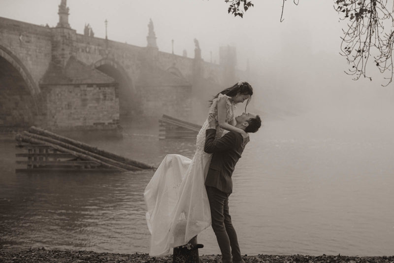 A monochrome image of a young man picking up his stylishly dressed bride against the backdrop of the Charles Bridge during their foggy Prague photoshoot.