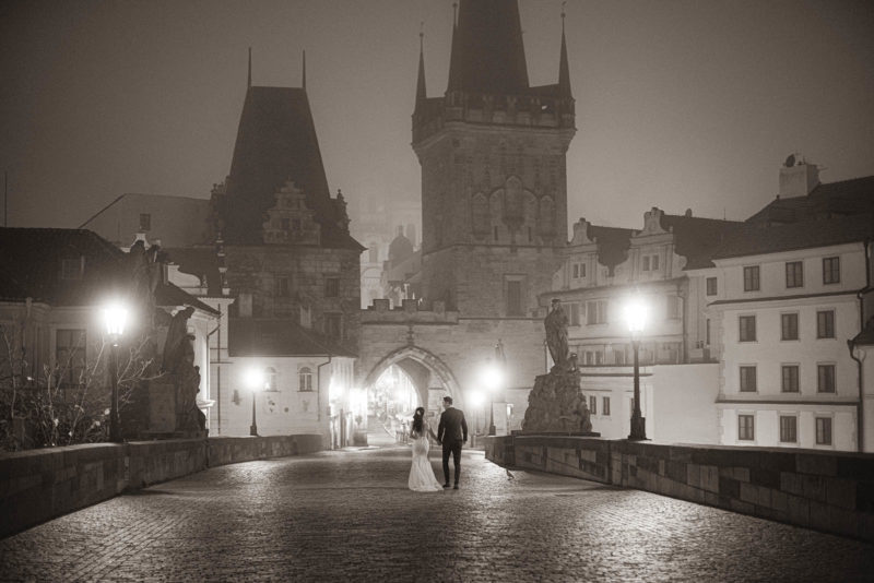 A stylishly dressed couple walk atop the gas lamp lit Charles Bridge as Prague is engulfed in a thick fog during their pre-wedding.