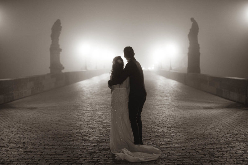 A monochrome image of a stylishly dressed couple embracing as they stand atop a fog shrouded Charles Bridge at night.