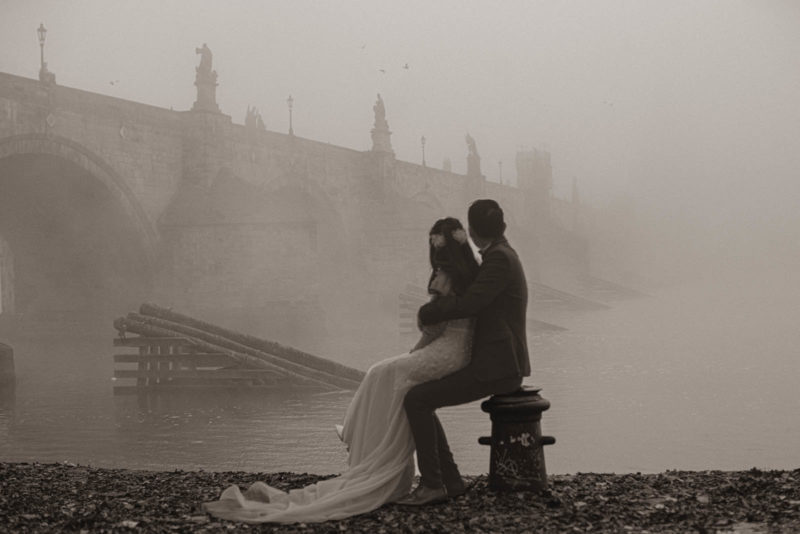 A monochrome image of a stylishly dressed couple wathching the mist surrounding the Charles Bridge during their foggy Prague photo session.