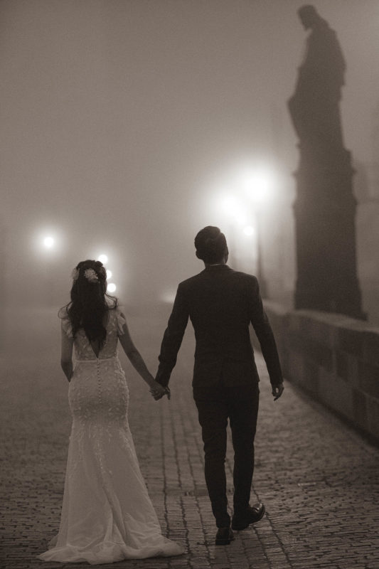 A monochrome image of a young, elegantly dressed couple walking hand-in-hand atop the night time scenery of a foggy Charles Bridge.