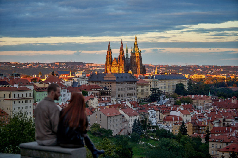 The stunning view of Prague Castle as the evening sun begins to fade against its historic facade as Max and Bryanna look on.